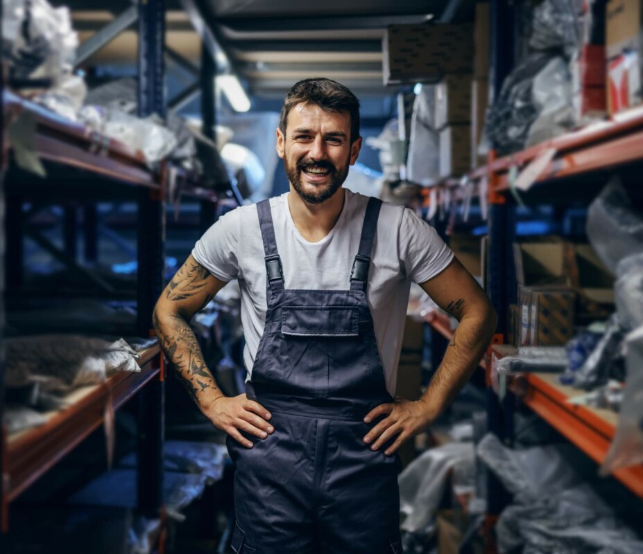 Smiling tattooed bearded worker in overalls standing in storage